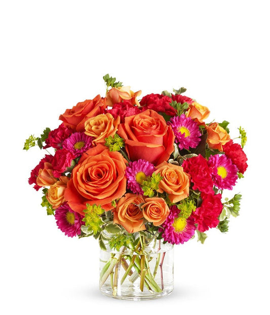 Floral arrangement featuring light orange roses, orange spray roses, hot pink carnations, and matsumoto asters in a clear glass vase.