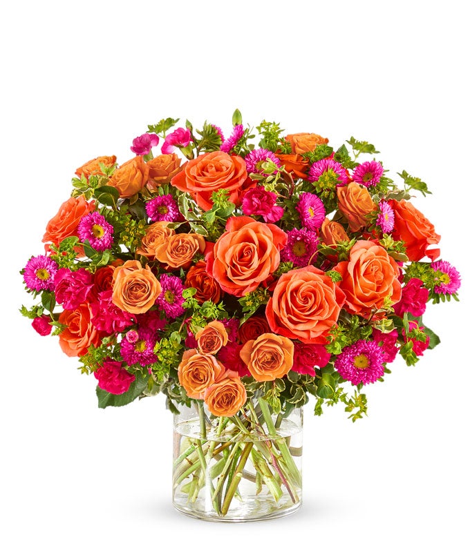 Floral arrangement featuring light orange roses, orange spray roses, hot pink carnations, and matsumoto asters in a clear glass vase.