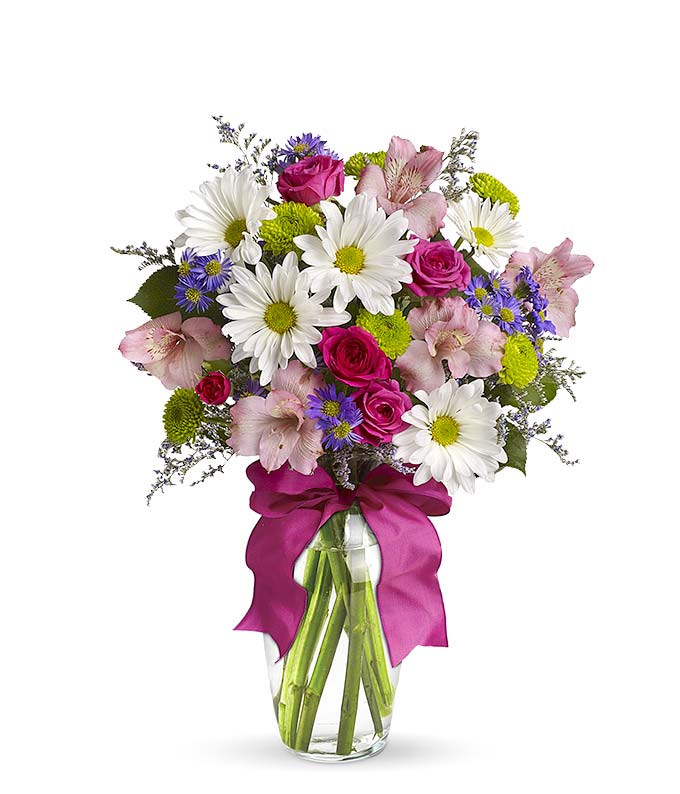 A cheerful floral arrangement featuring pink spray roses, white daisies, green button poms, purple Monte Casino, and limonium, all beautifully arranged in a glass vase with a hot pink ribbon. This vibrant mix offers a playful and lively display.