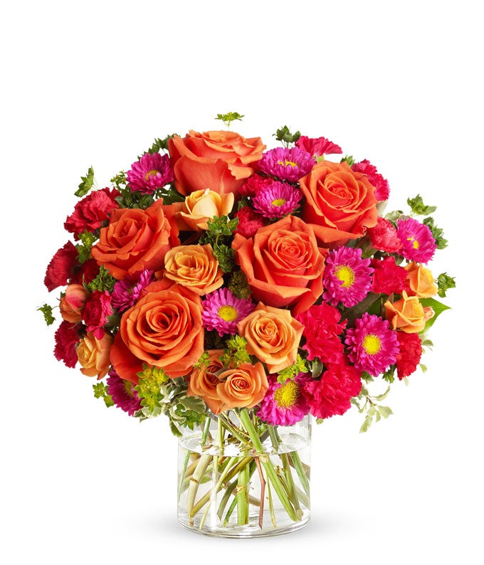 Floral arrangement featuring light orange roses, orange spray roses, hot pink carnations, and matsumoto asters in a clear glass vase.