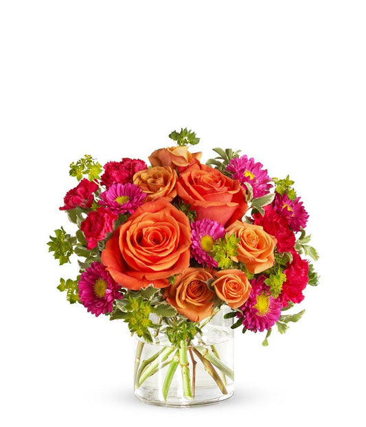 Floral arrangement featuring light orange roses, orange spray roses, hot pink carnations, and matsumoto asters in a clear glass vase.