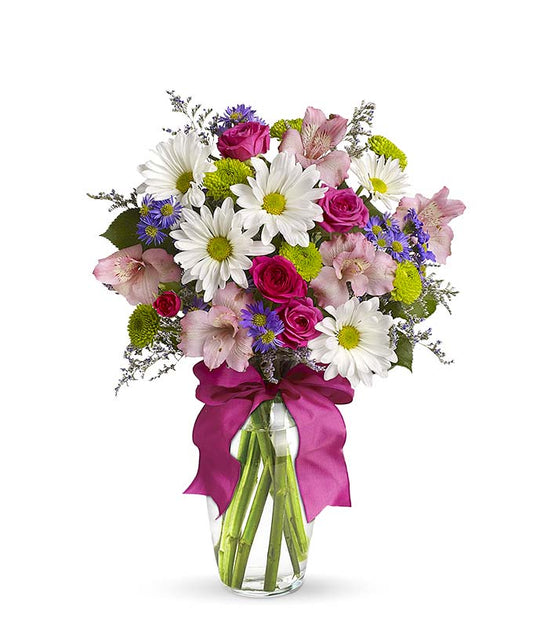 A cheerful floral arrangement featuring pink spray roses, white daisies, green button poms, purple Monte Casino, and limonium, all beautifully arranged in a glass vase with a hot pink ribbon. This vibrant mix offers a playful and lively display.