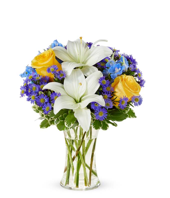 Yellow roses, blue delphinium and white lilies in a clear glass vase