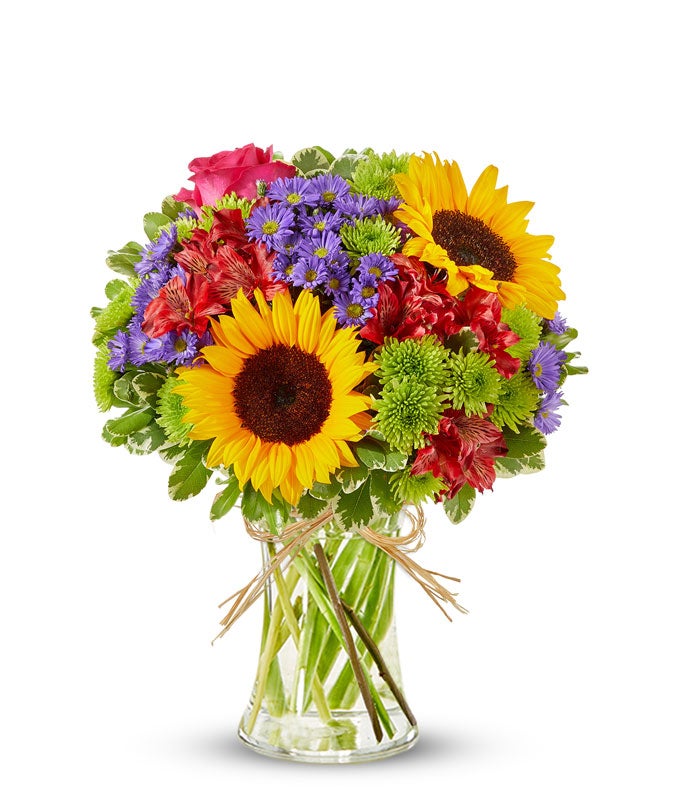 Bright mixed flower bouquet featuring yellow sunflowers, hot pink roses, red alstroemeria, green button poms, and purple asters in a clear glass vase tied with raffia ribbon.
