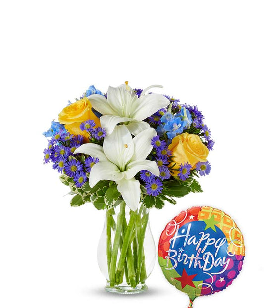 Birthday flower bouquet with white lilies, yellow roses, blue delphinium, and purple asters in a clear glass vase, paired with a colorful Happy Birthday balloon.